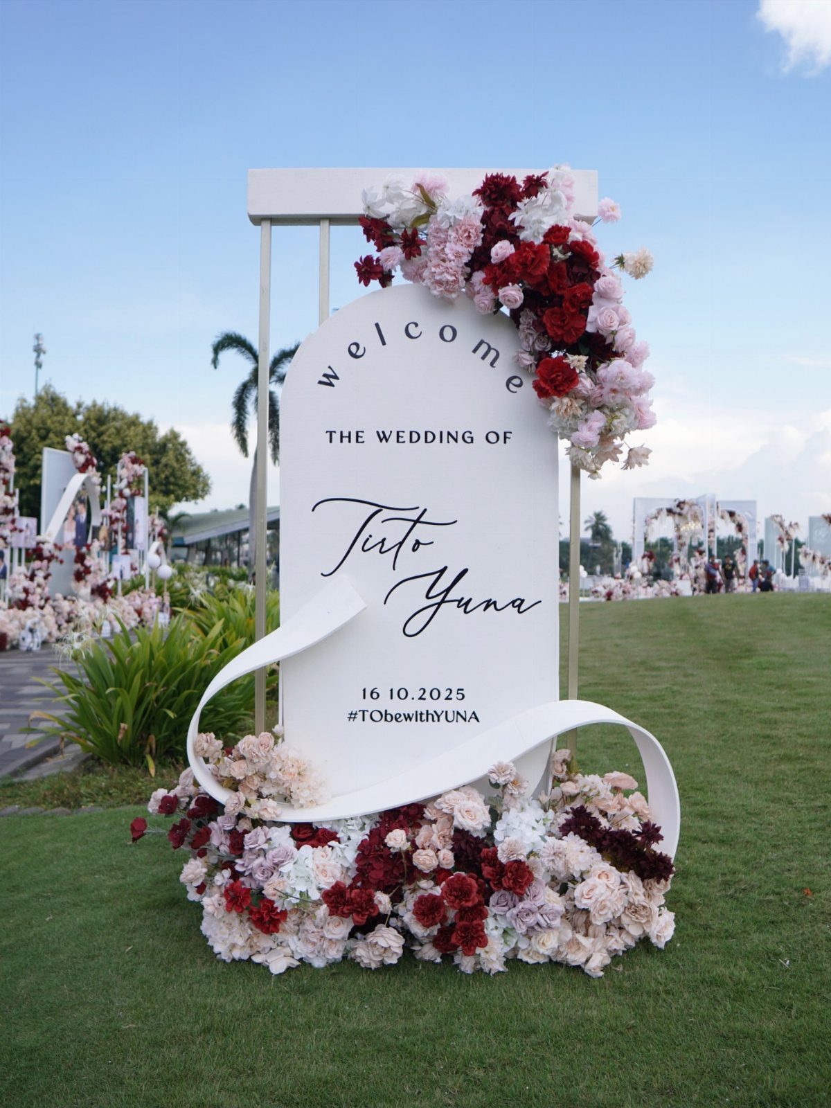 A white oval wedding welcome sign decorated with pink and red flowers on a green lawn.