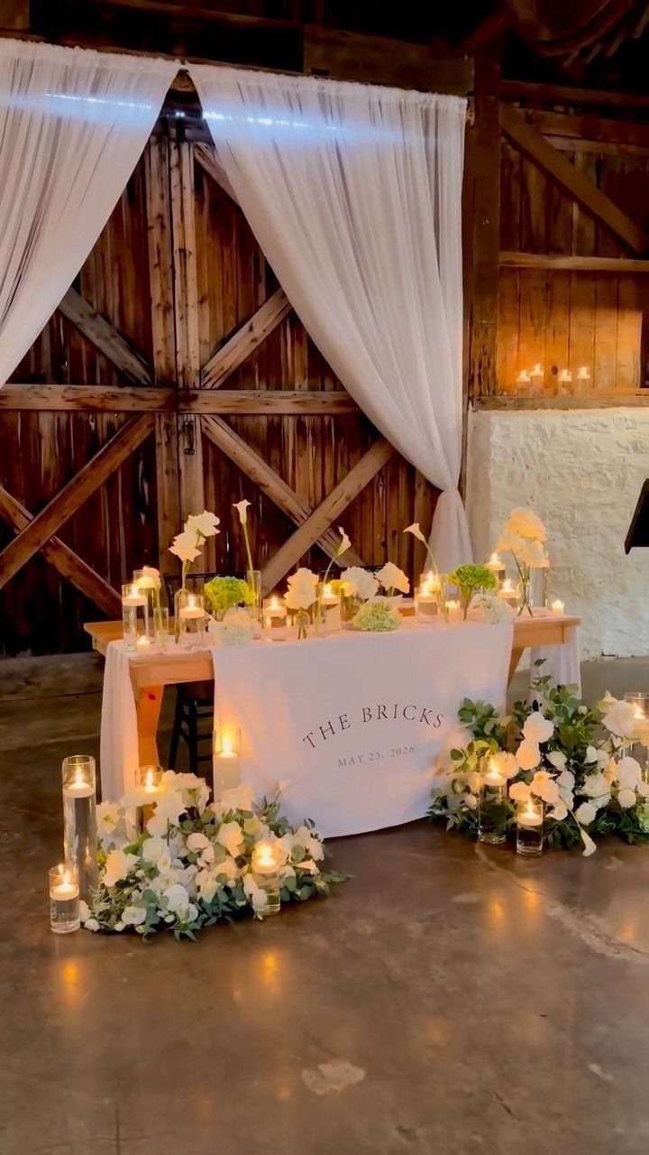A decorated wedding sweetheart table with white flowers and glowing candles inside a rustic wooden barn.