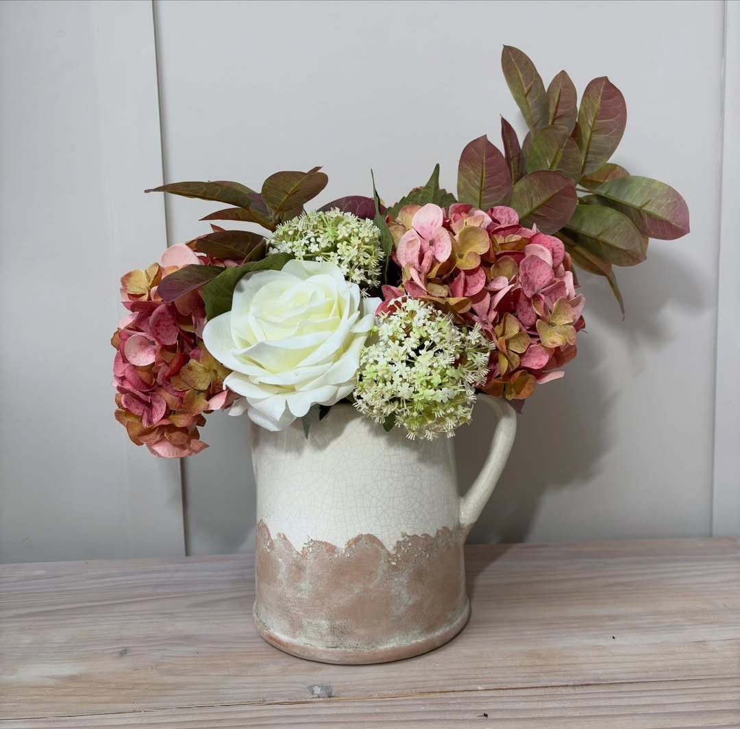 Floral arrangement with white and pink hydrangeas in a rustic ceramic pitcher on wood.