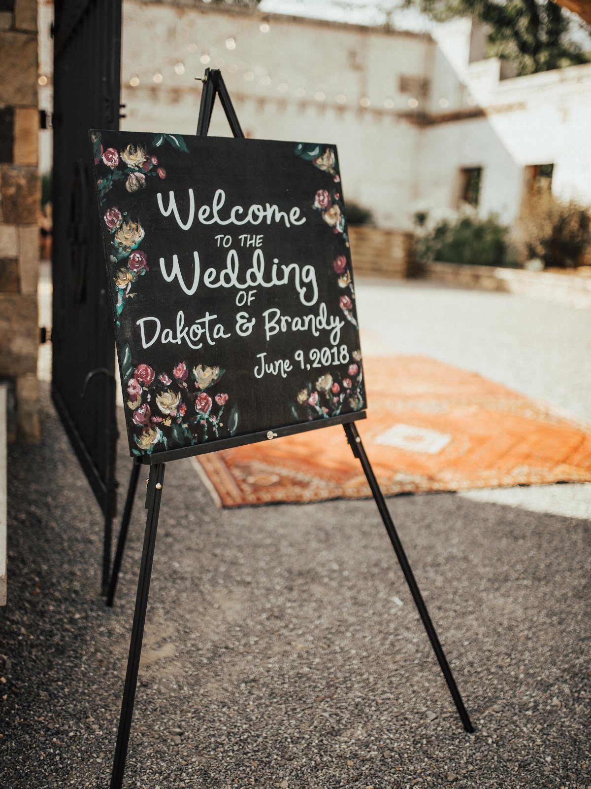 A chalkboard wedding welcome sign with floral decorations on a black easel outdoors.