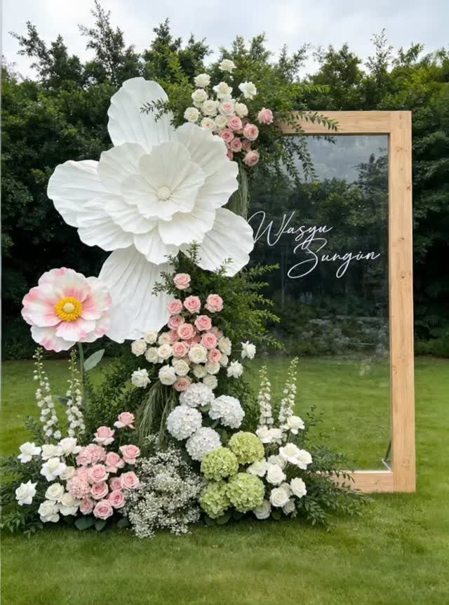Large white and pink floral arrangement decorating a wooden framed glass backdrop on grass.
