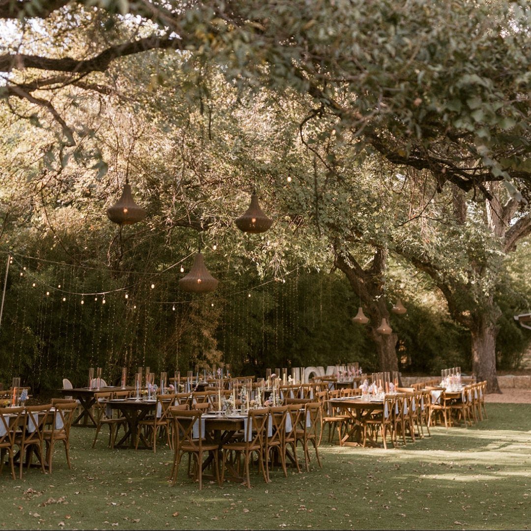 Bell-shaped lanterns hanging from large tree branches over an outdoor wedding reception setup.