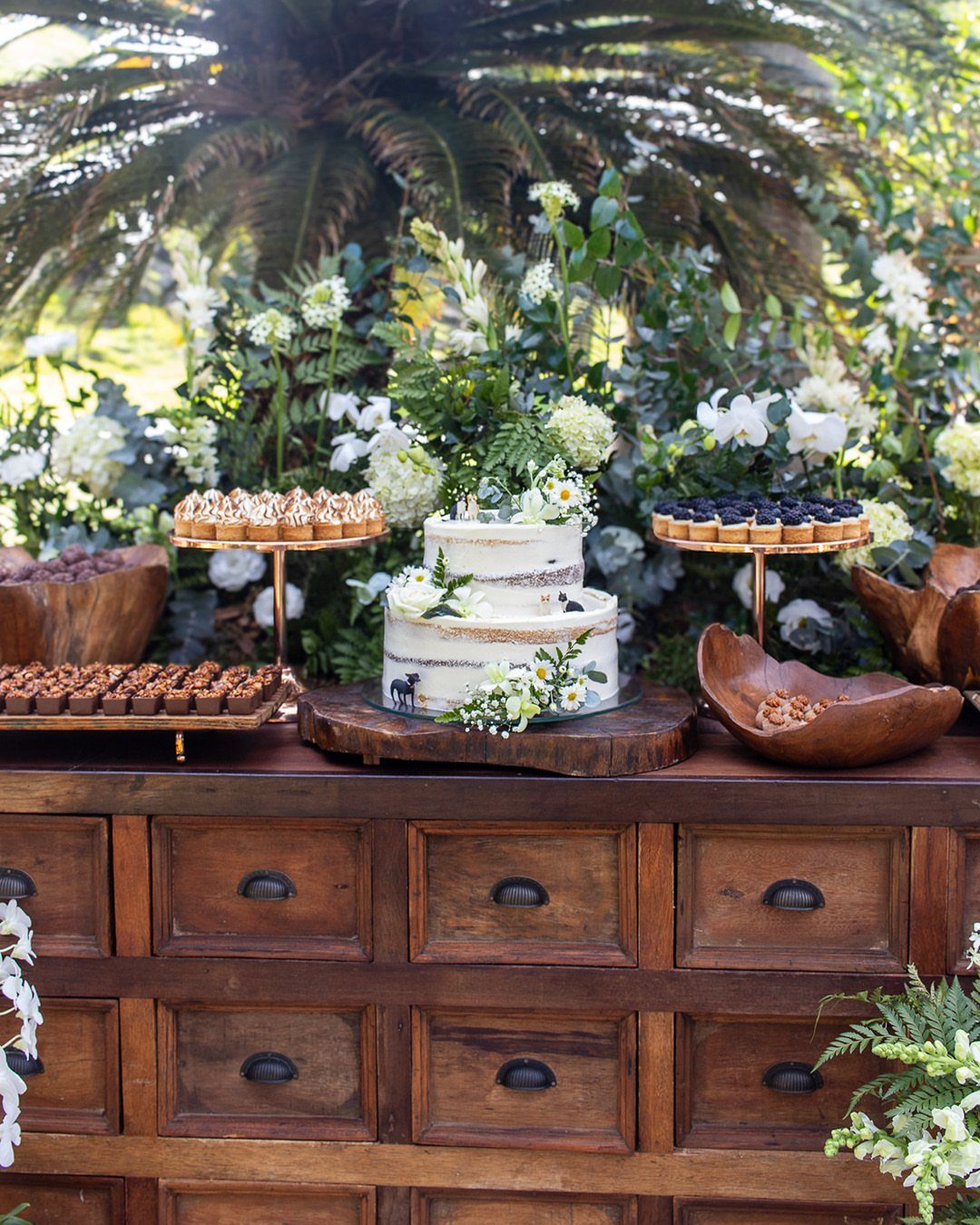A rustic wooden dresser displaying a white cake and various desserts amidst lush greenery.