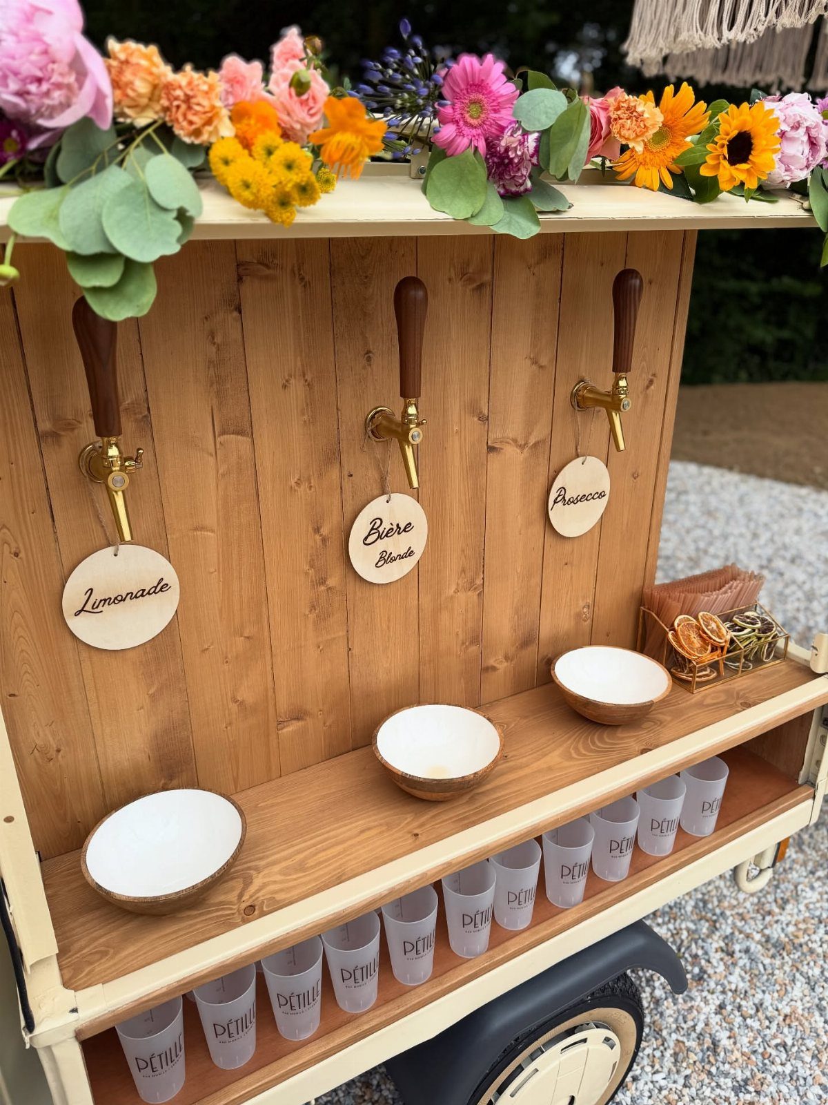 A wooden rustic drink cart with gold dispensers, floral decorations, and small bowls on a counter.