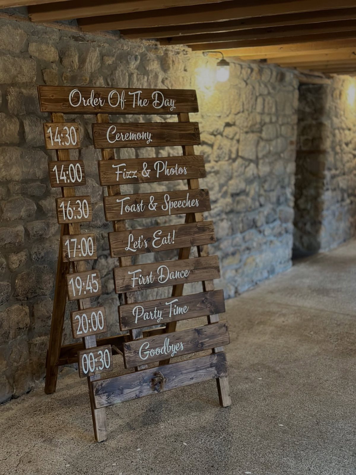 A rustic wooden wedding schedule sign leaning against a stone wall in a dimly lit room.
