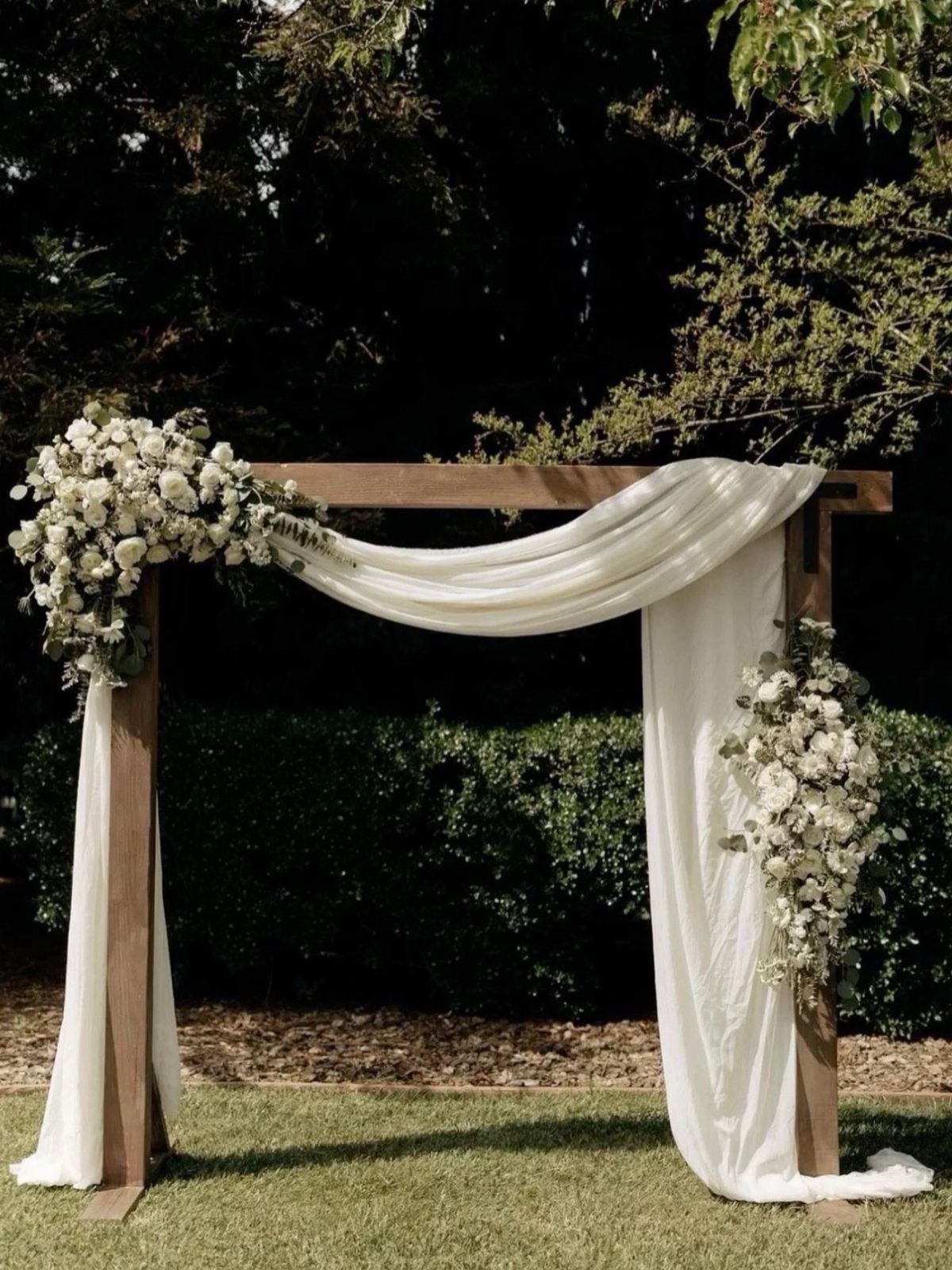 Simple Garden Wedding Reception Outdoor: 14 Stylish Ideas to Copy 6 A wooden wedding arbor adorned with white flowers and draped white fabric in a garden.