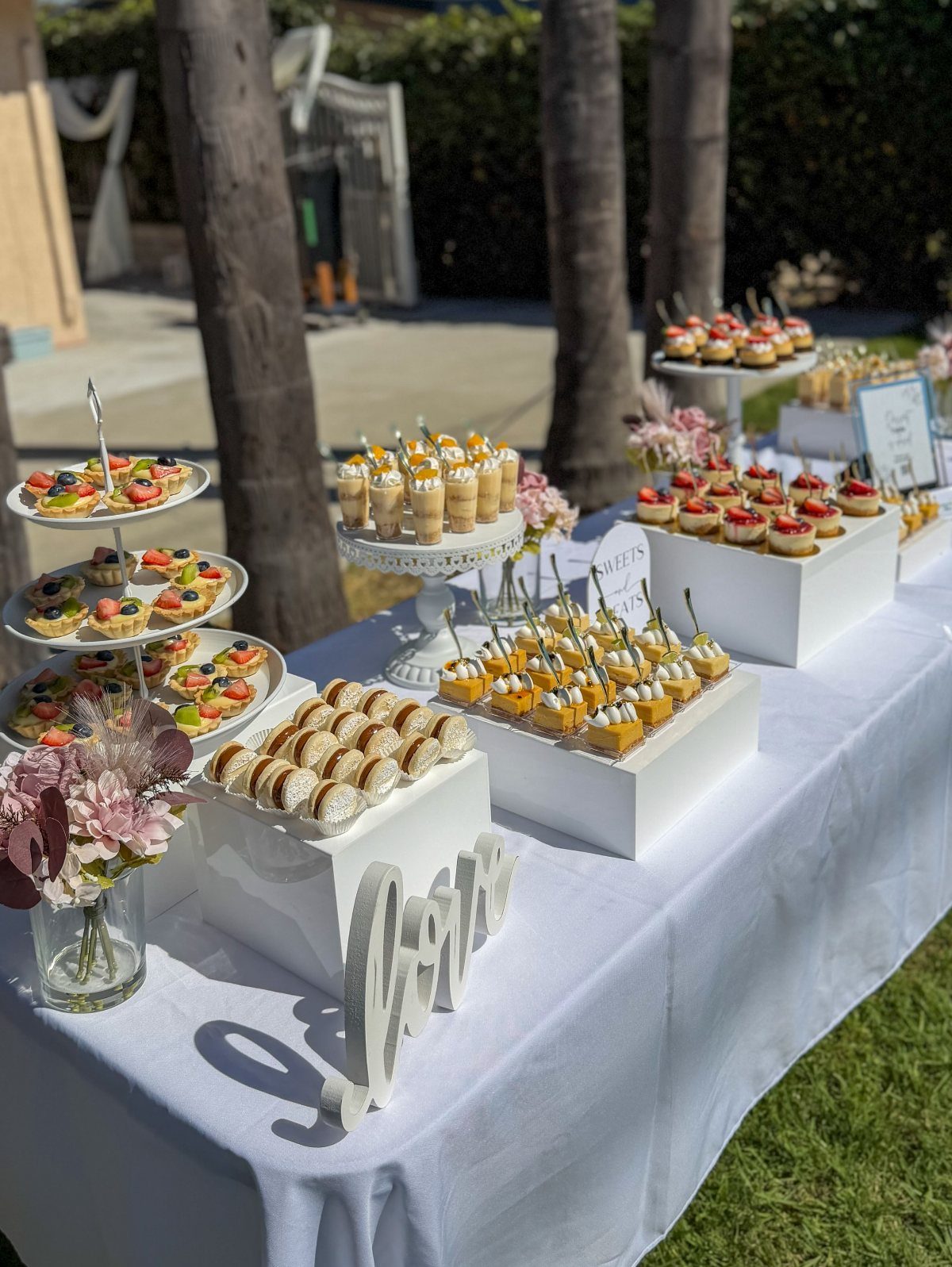 Simple Garden Wedding Reception Outdoor: 14 Stylish Ideas to Copy 7 Various mini fruit tarts and small pastries arranged on a white dessert table outdoors.
