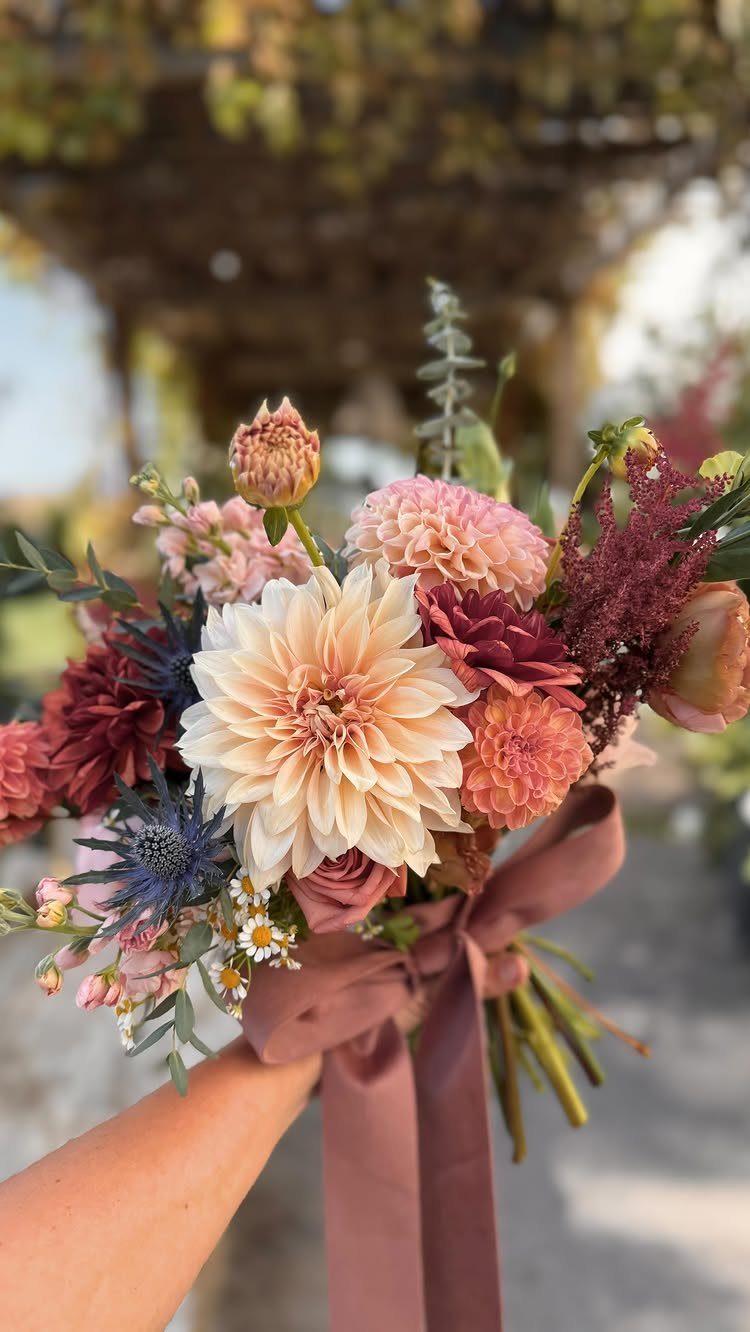 Simple Garden Wedding Reception Outdoor: 14 Stylish Ideas to Copy 2 A hand holding a seasonal bouquet of dahlias, eryngium, and daisies with a mauve ribbon.
