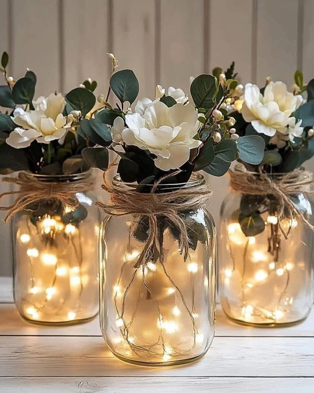 8 Simple Wedding Centerpieces for Budget-Friendly Tables 1 Three mason jars containing white flowers, greenery, and glowing fairy lights on a white surface.