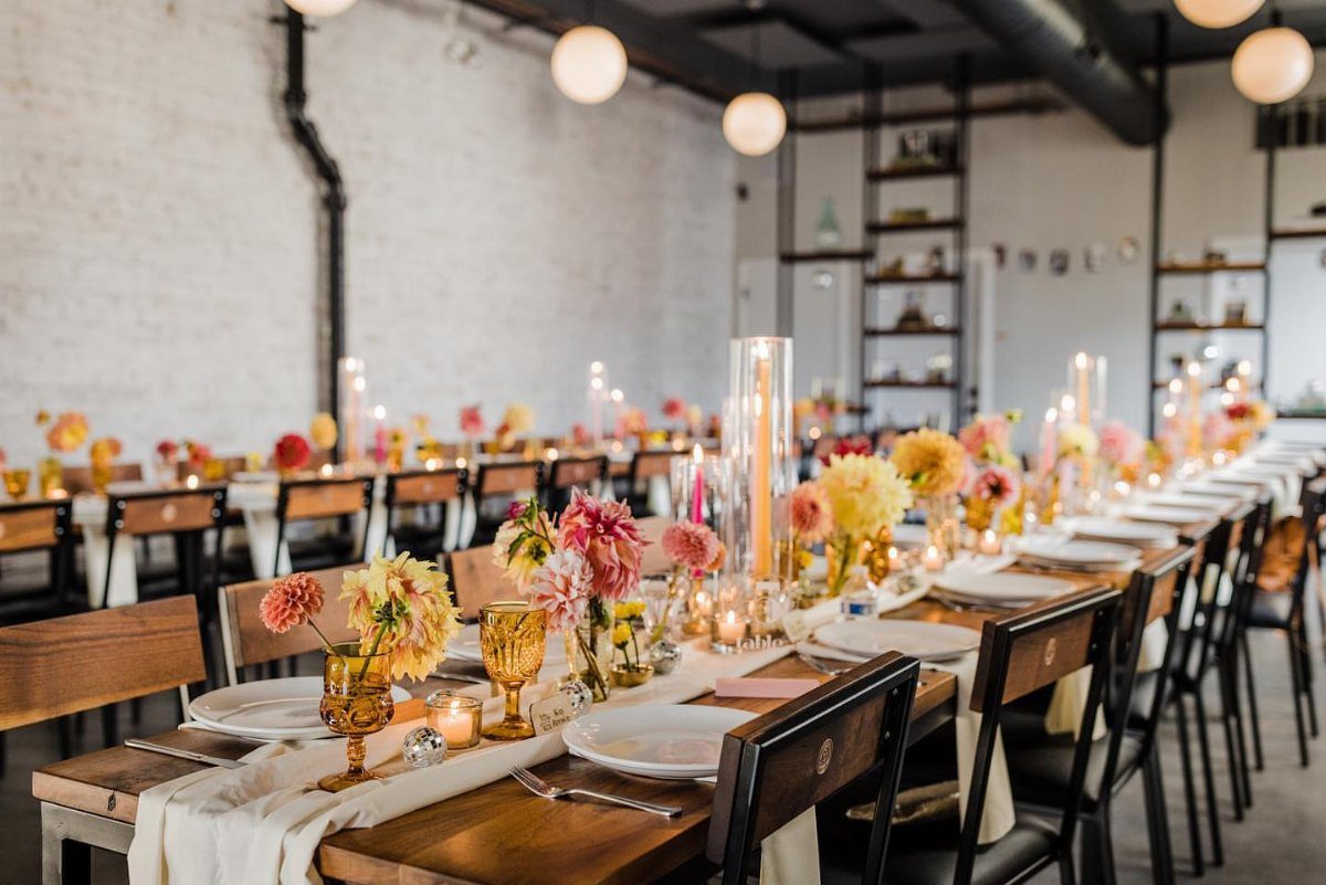 11 Thrifted Wedding Decor Ideas to Steal for Real Budgets 9 Long wooden wedding tables decorated with amber glassware, colorful flowers, and tall candles in an industrial space.