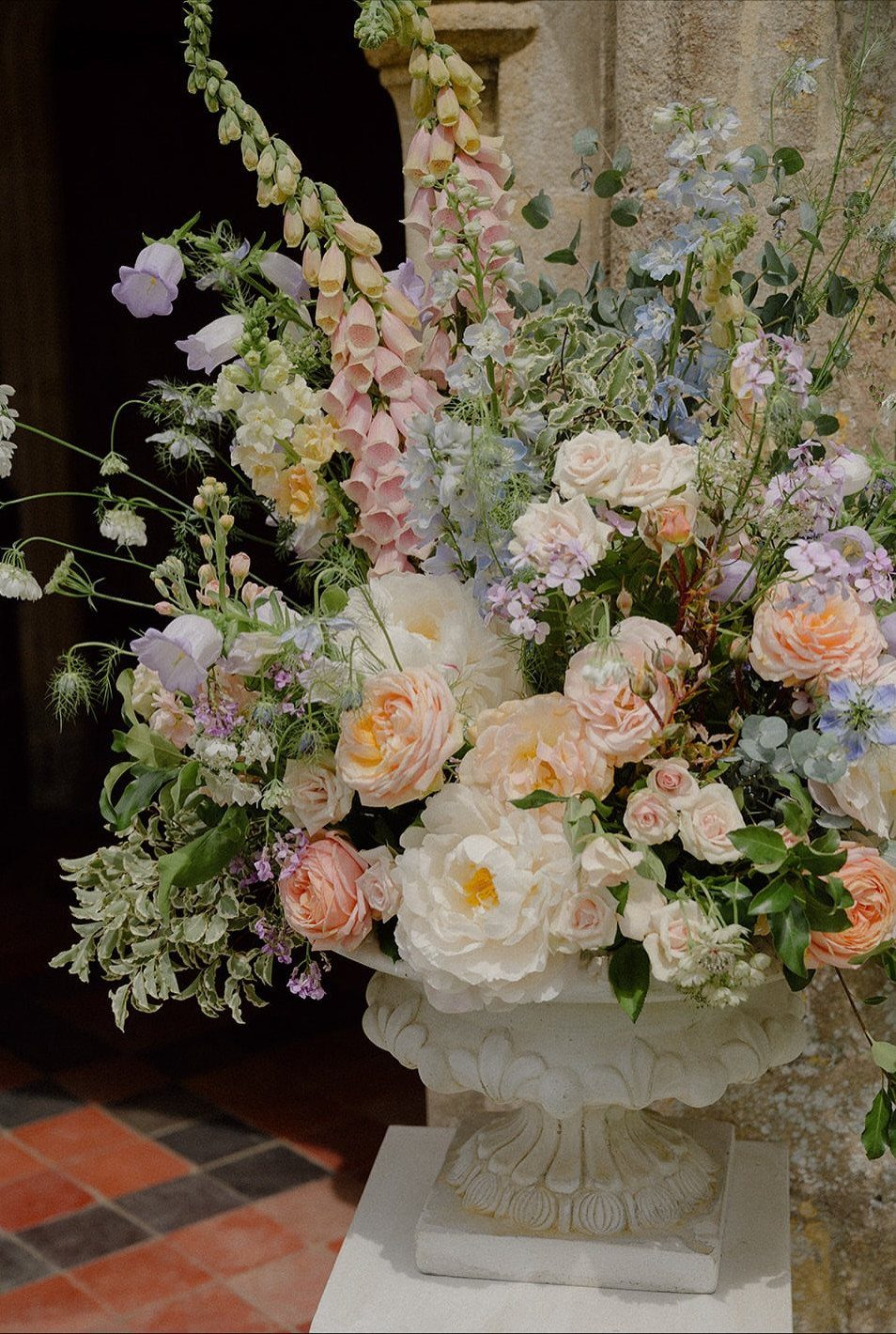 A large white stone urn filled with pastel roses and wildflowers in a garden setting.