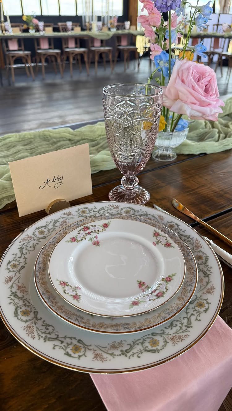 Vintage floral china plates and a pink patterned glass set on a wooden wedding reception table.