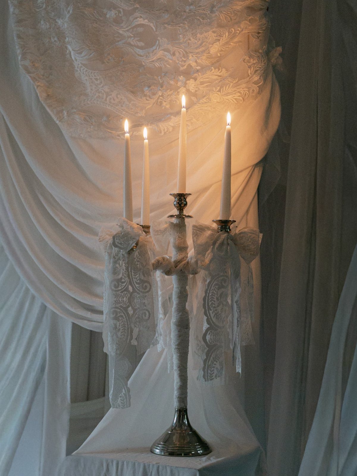 Silver candelabra with lit white candles and lace ribbons against draped white fabric.