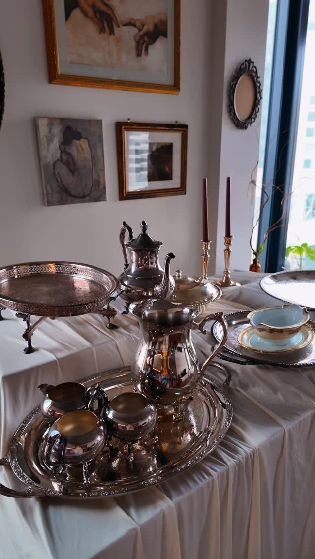 Ornate silver tea sets and serving trays arranged on a white tablecloth in a room.