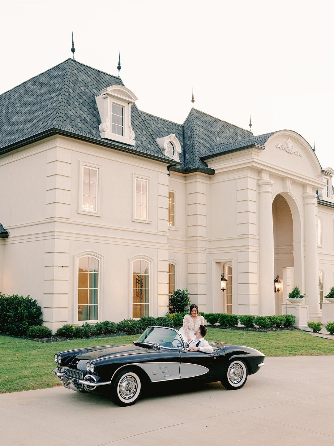 A black vintage convertible car parked on a driveway in front of a large white mansion.