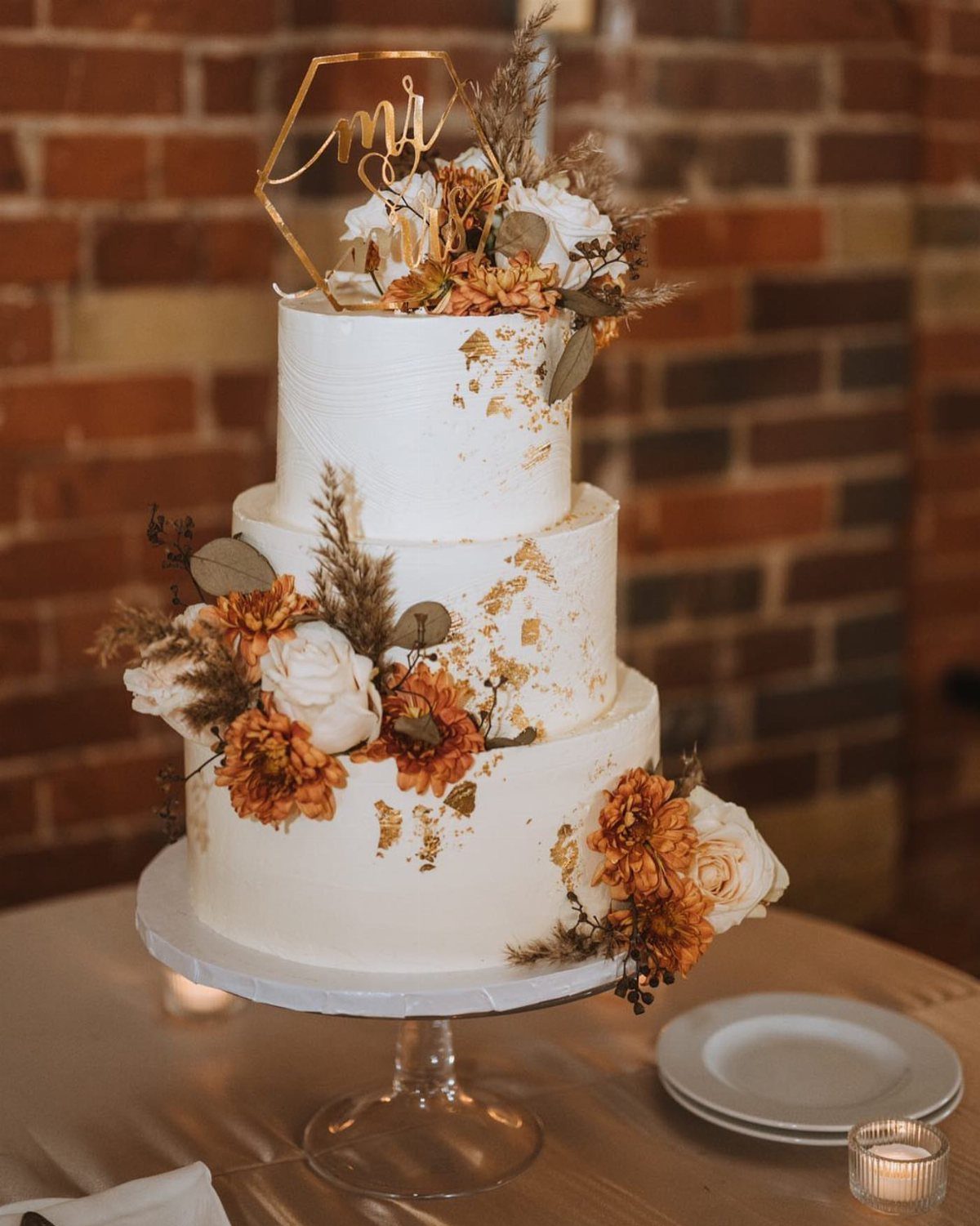 Three-tier white buttercream wedding cake decorated with gold leaf and dried flowers on a glass stand.