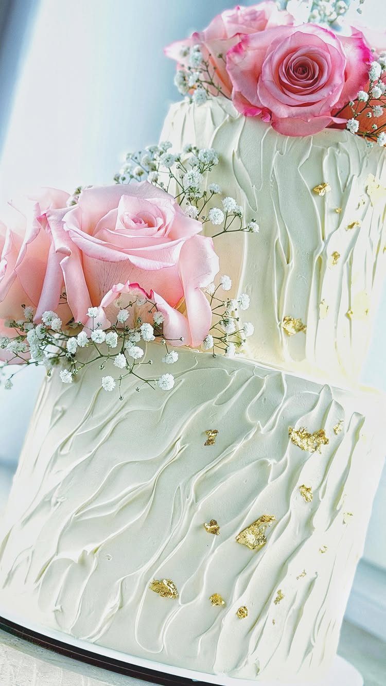 Two-tier white wedding cake with textured buttercream, gold leaf, and pink roses.