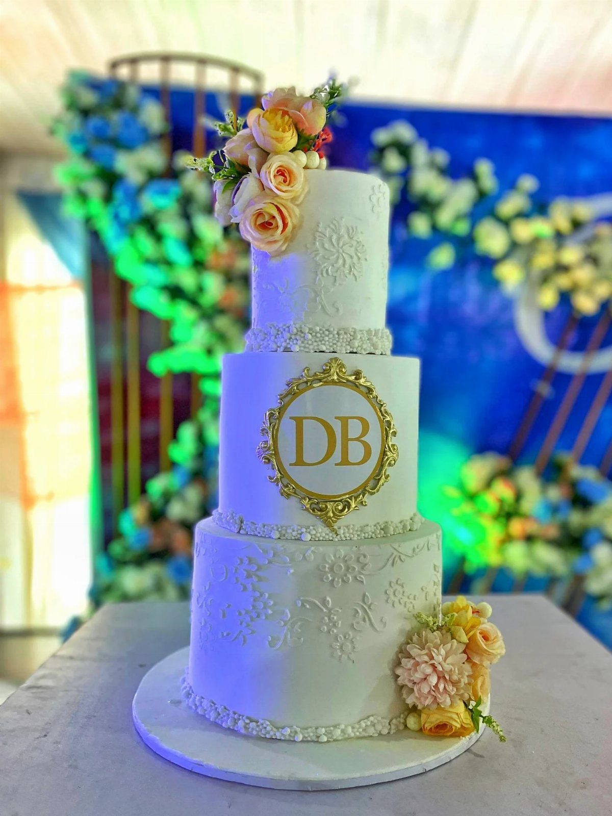 A three-tier white wedding cake with gold monogram and peach roses on a floral background.
