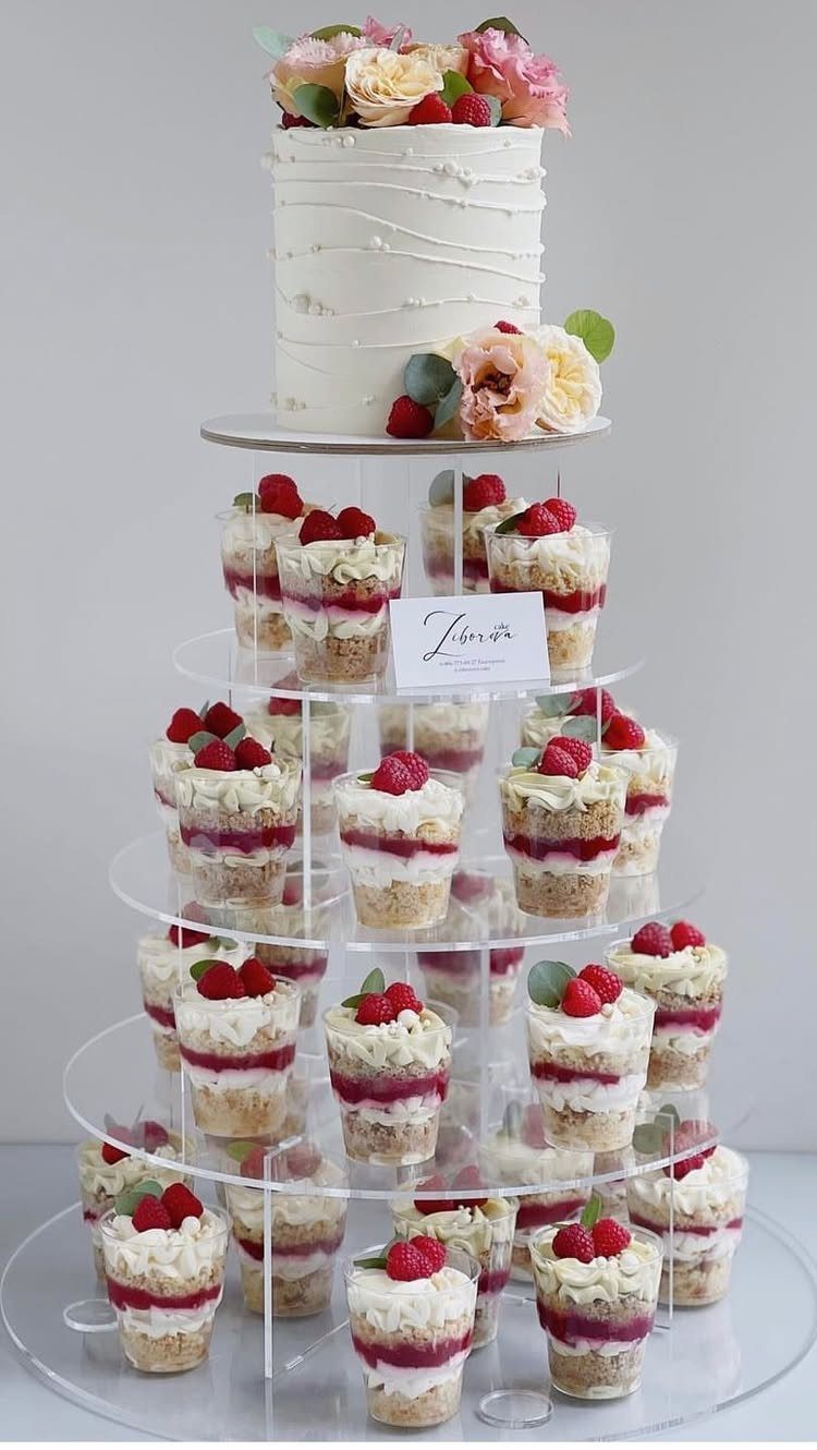 How to Choose a Wedding Cake Stand: 8 Styles for Every Reception 4 A clear acrylic tiered stand displaying a white floral cake and many small dessert cups.