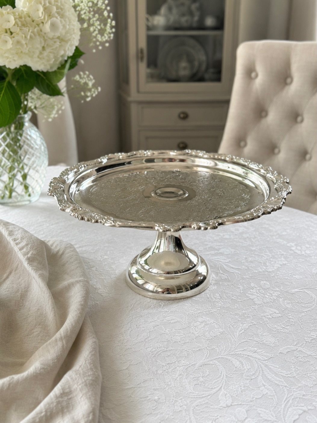 How to Choose a Wedding Cake Stand: 8 Styles for Every Reception 5 A silver metal cake stand with scalloped edges on a white lace tablecloth.