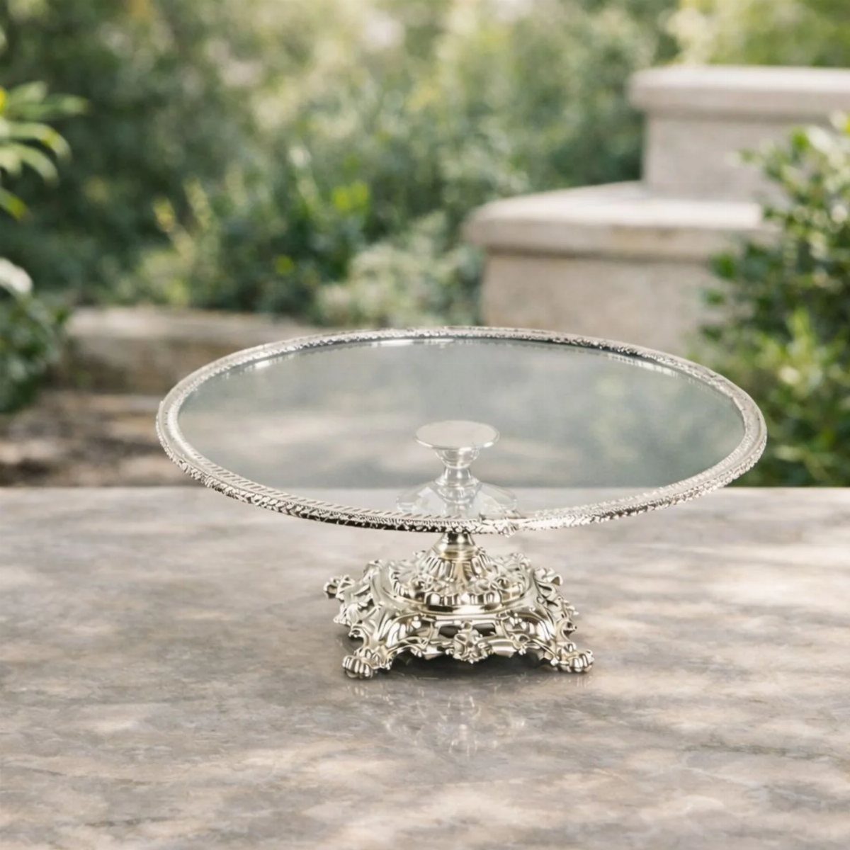How to Choose a Wedding Cake Stand: 8 Styles for Every Reception 8 An ornate silver pedestal cake stand with a glass plate on a marble table outdoors.