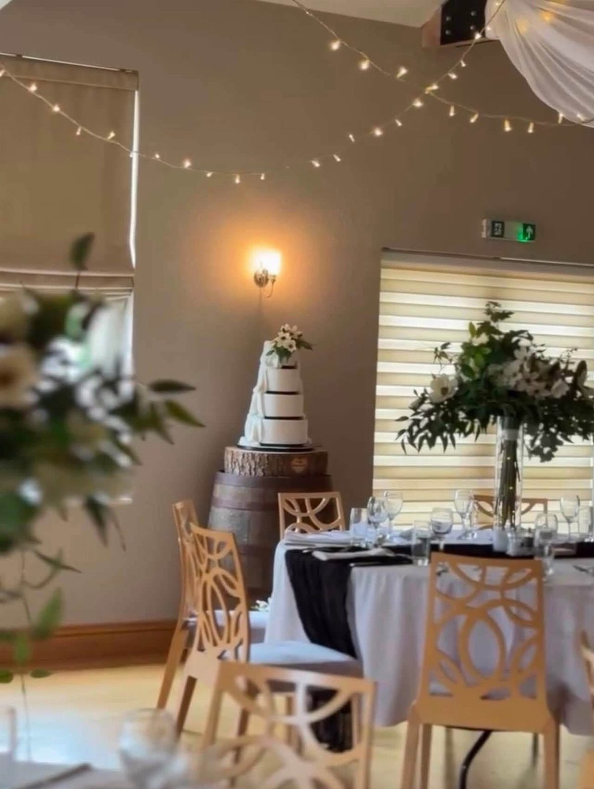 How to Choose a Wedding Cake Stand: 8 Styles for Every Reception 7 A white tiered wedding cake displayed on a wooden barrel in a decorated reception room.