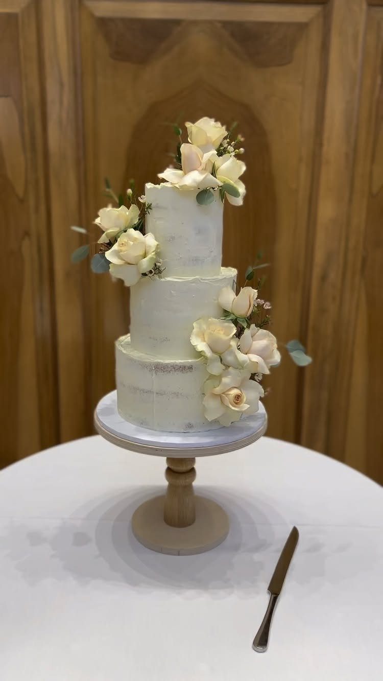 How to Choose a Wedding Cake Stand: 8 Styles for Every Reception 3 A three-tier white wedding cake with cream roses on a rustic wooden cake stand.