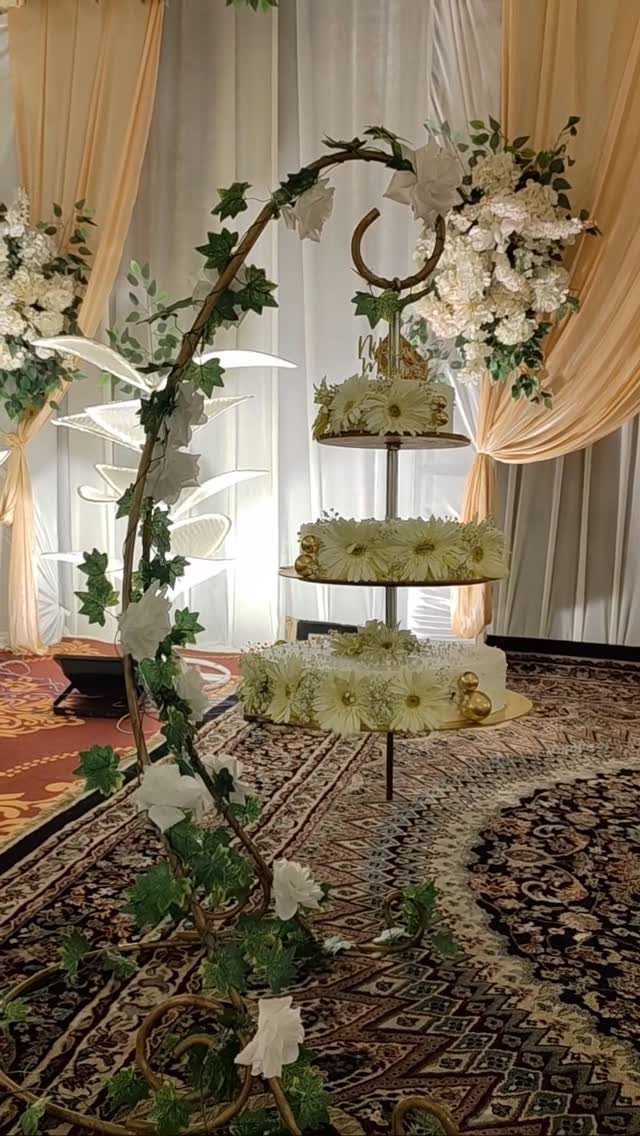 How to Choose a Wedding Cake Stand: 8 Styles for Every Reception 2 A three-tier wedding cake stand decorated with white flowers on a patterned rug.