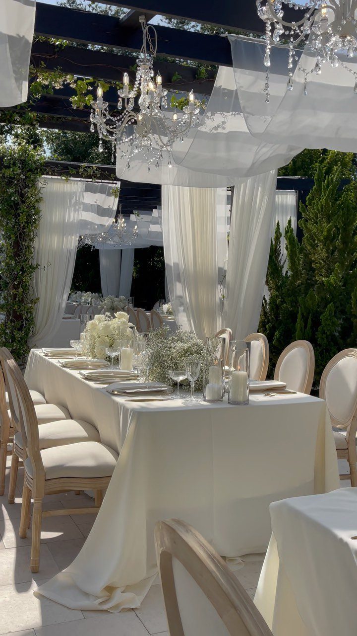 White wedding table setting with floral centerpieces and crystal chandeliers under white draped fabric.