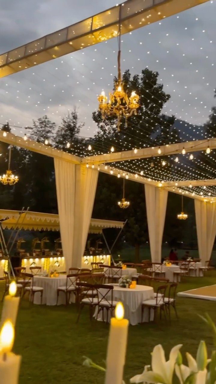 Twinkling fairy lights and golden chandeliers hang over an outdoor wedding reception with white draped fabric.