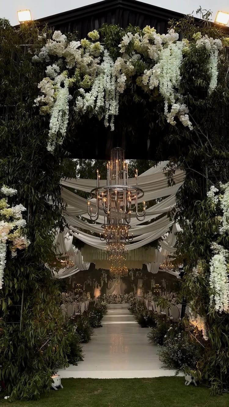 A white floral archway framing an elegant outdoor wedding reception with draped fabric and chandeliers.