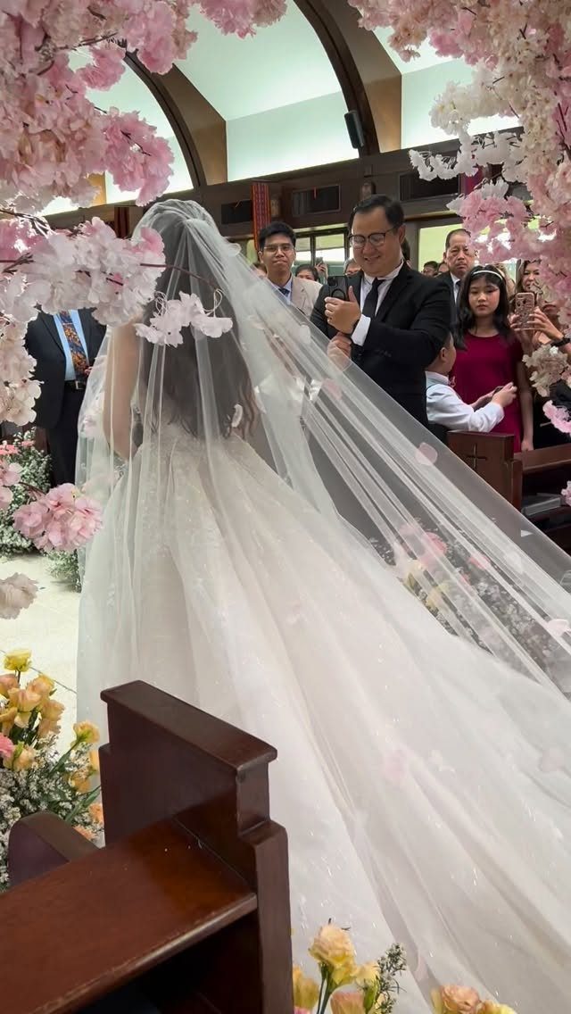 A bride in a white gown walks through pink cherry blossom floral decorations in a church.