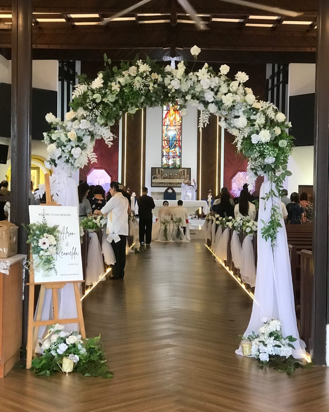 A white floral arch with greenery stands at the entrance of a church aisle leading to the altar.