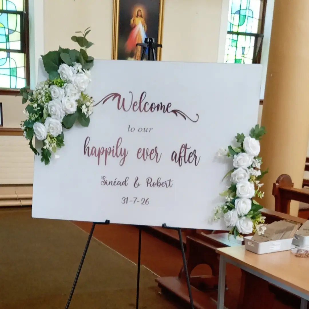 A white wedding welcome sign with floral decorations stands on an easel inside a church.