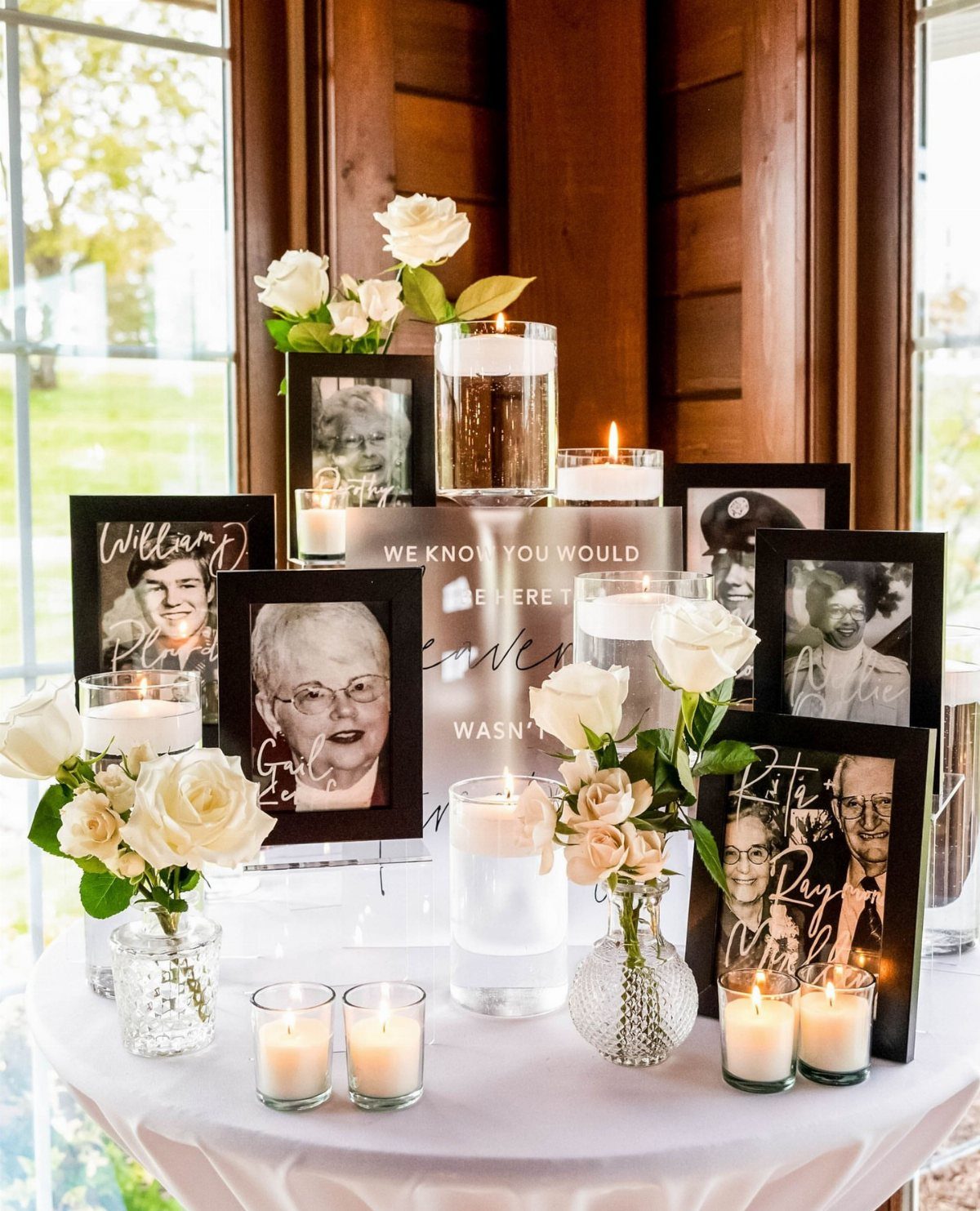 Framed black and white portraits surrounded by white roses and lit candles on a memorial table.