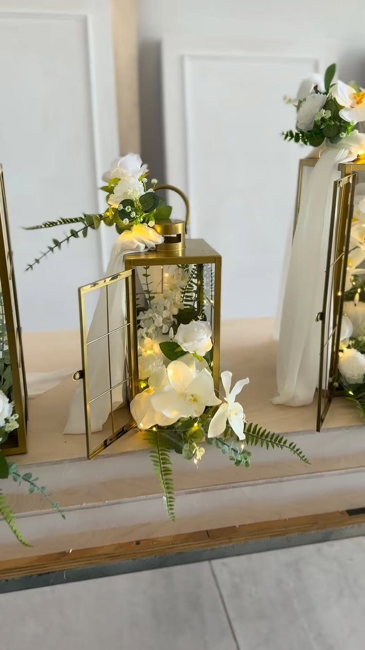 20 Wedding Decor DIY Ideas for Ceremony and Reception 8 Gold metal lanterns decorated with white flowers and flowing white fabric on a wooden ledge.