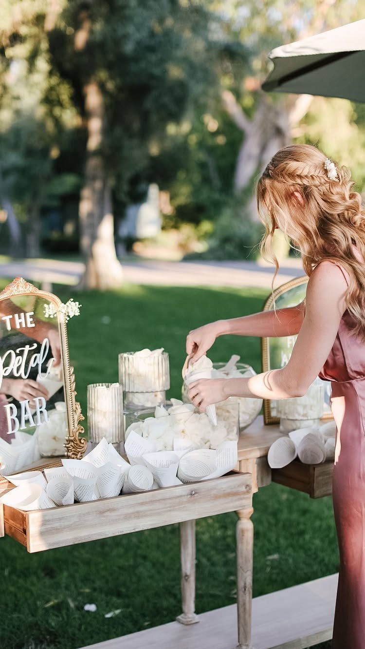 22 Wedding Decor Garden Theme Ideas for Every Reception Style 21 A woman arranging white treats at an outdoor wooden table labeled The Petal Bar.