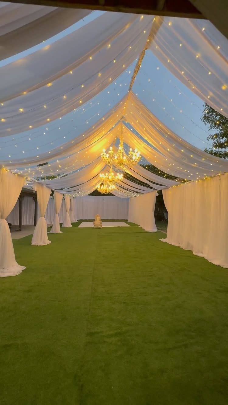 22 Wedding Decor Garden Theme Ideas for Every Reception Style 18 White fabric canopy with twinkle lights and chandeliers hanging over a green lawn at twilight.