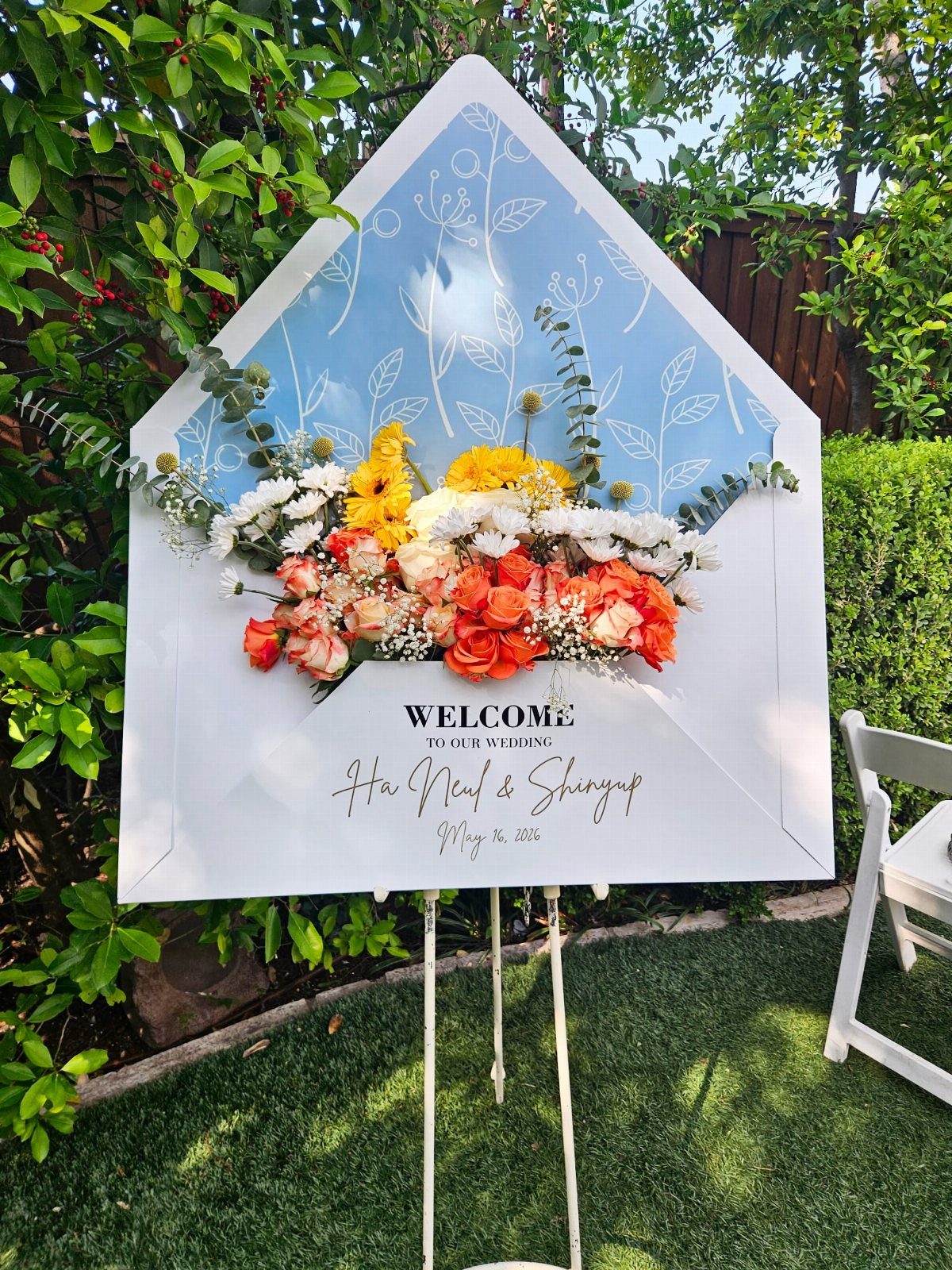22 Wedding Decor Garden Theme Ideas for Every Reception Style 7 An envelope-shaped wedding welcome sign decorated with colorful flowers on a white easel outdoors.
