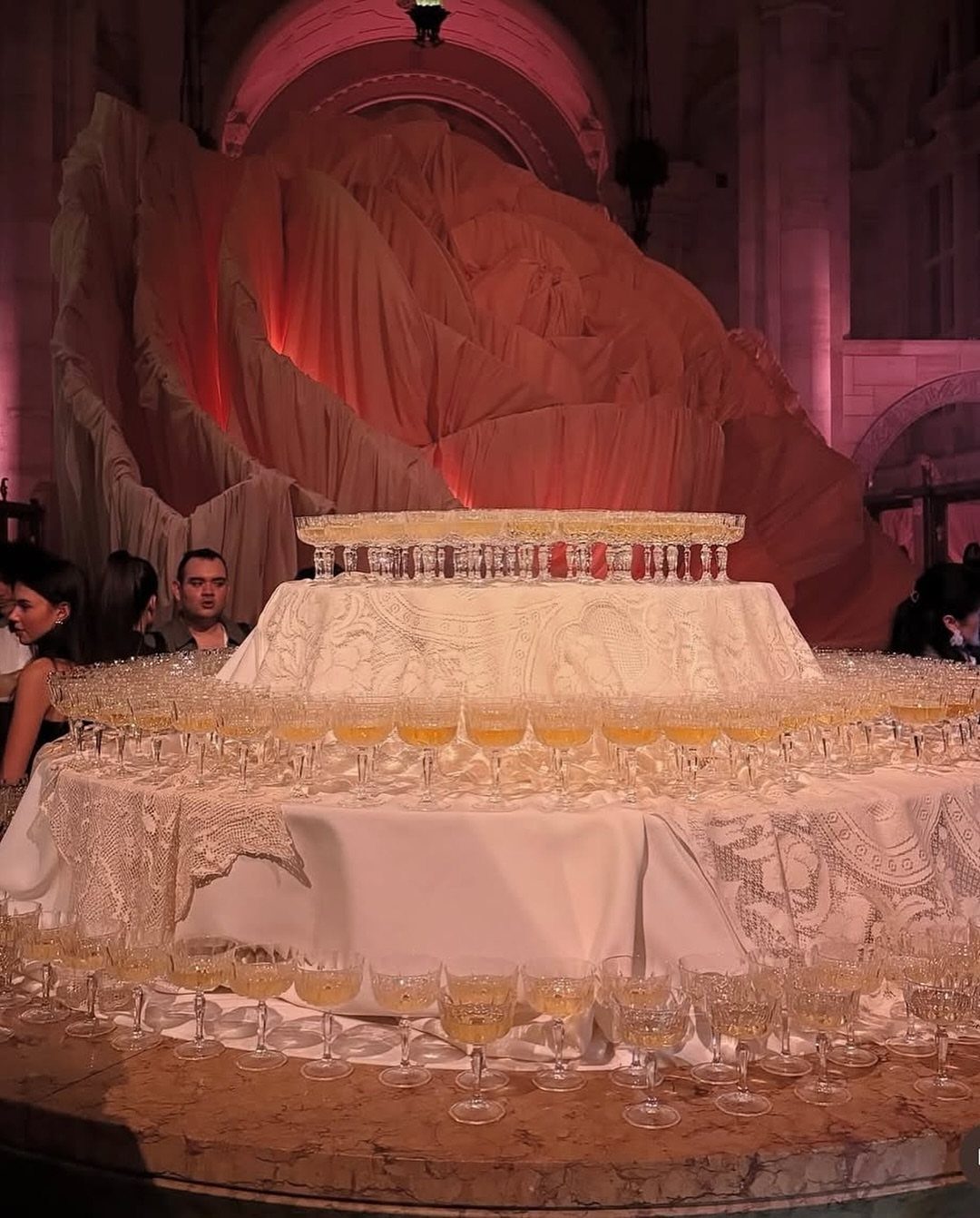 Hundreds of champagne glasses arranged on a tiered lace-covered table with draped fabric in the background.
