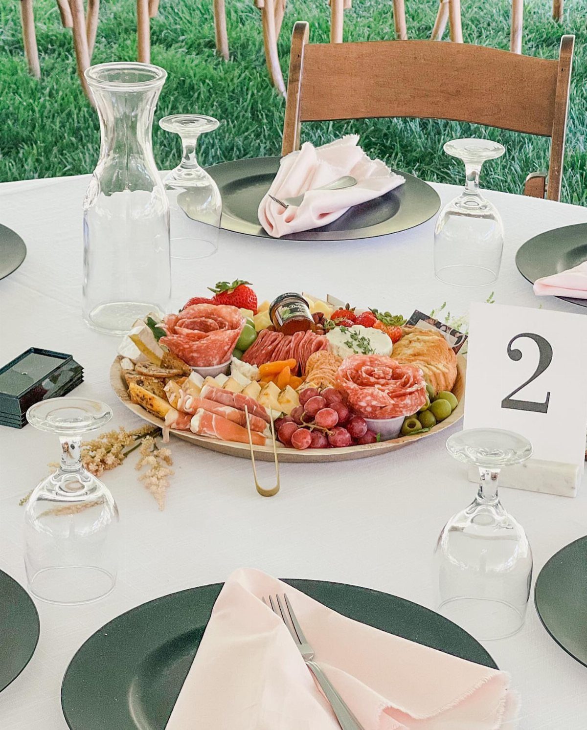 A decorated wedding table featuring a large charcuterie platter centerpiece with meats, cheeses, and fruit.