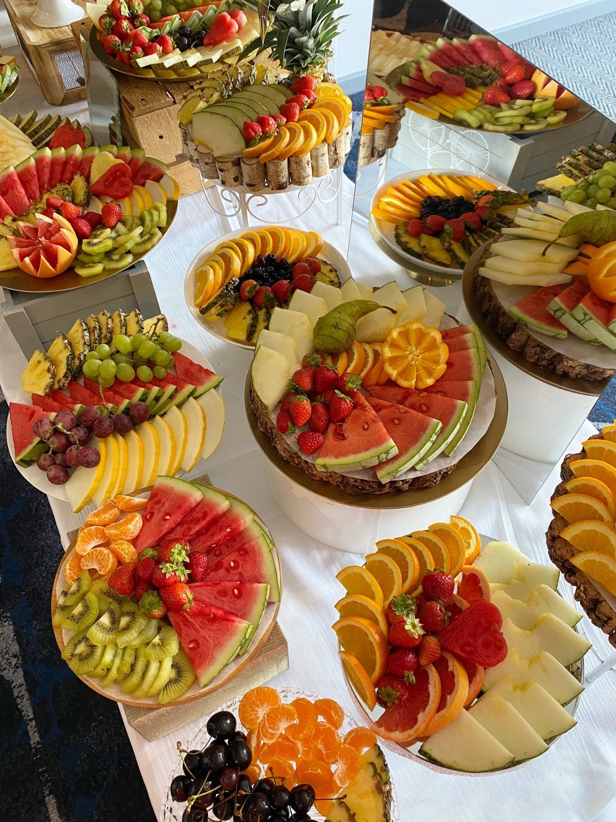 Assorted platters of sliced watermelon, kiwi, oranges, and strawberries arranged on a white table.