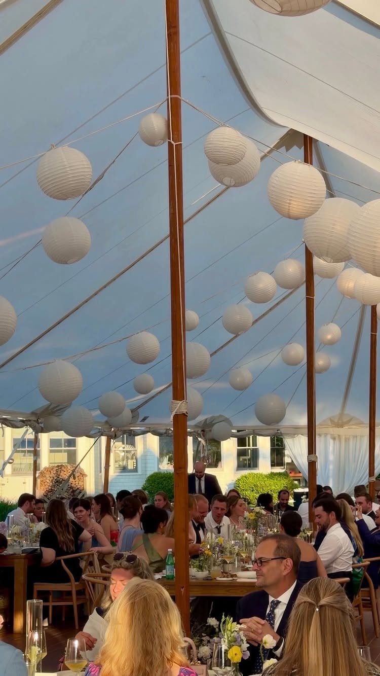 White paper lanterns hanging from the ceiling of a large white event tent over wedding guests.