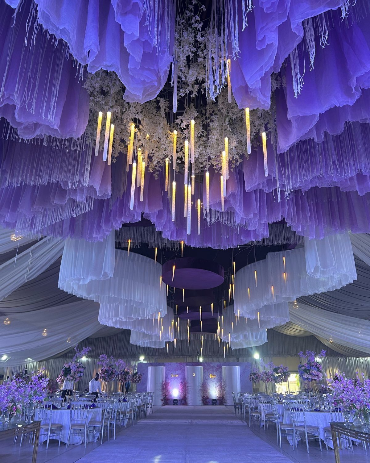 11 Wedding Decor Room Ideas for Your Reception in 2026 6 Purple and white fabric drapes hanging from a ceiling with long glowing vertical lights in a ballroom.