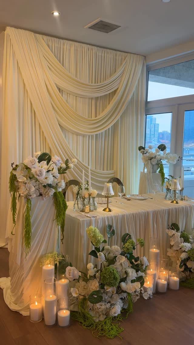 11 Wedding Decor Room Ideas for Your Reception in 2026 3 A decorated sweetheart table featuring white flowers, greenery, and glowing pillar candles against draped fabric.