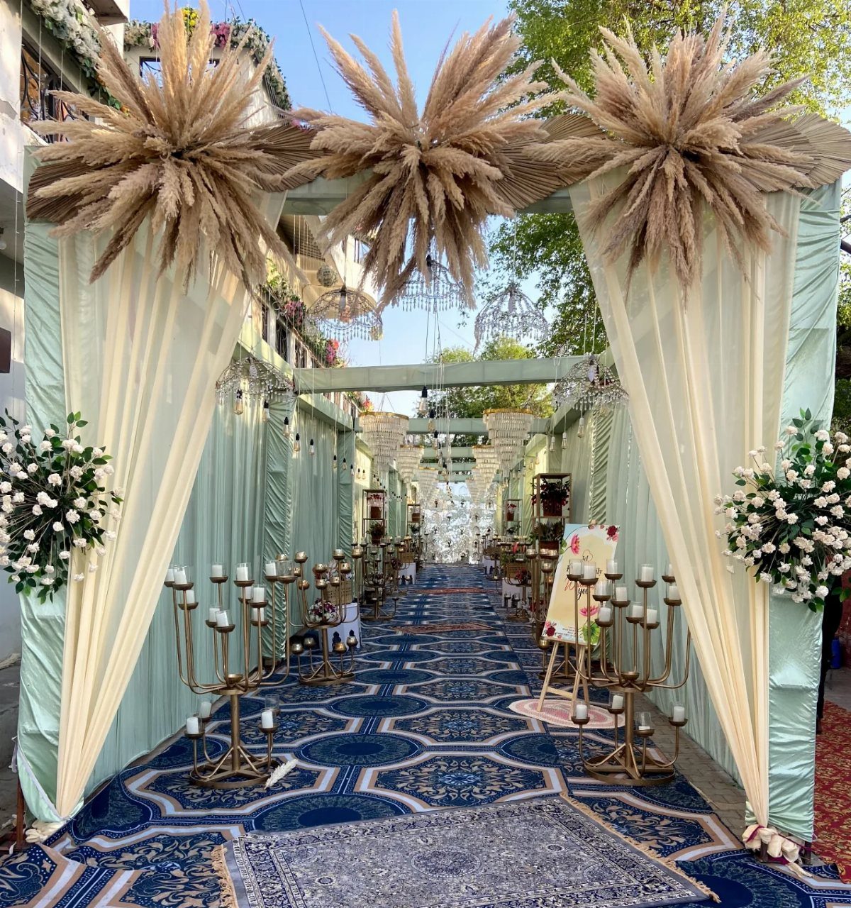13 Wedding Decor Tent Ideas for 2026 Receptions 5 Decorated wedding entrance with pampas grass, draped fabric, gold candelabras, and a blue patterned carpet aisle.