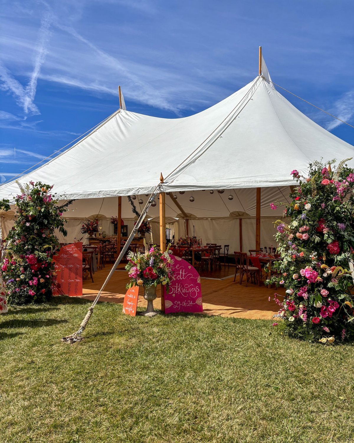 13 Wedding Decor Tent Ideas for 2026 Receptions 8 A large white sailcloth pole tent decorated with vibrant pink and red floral arrangements on green grass.