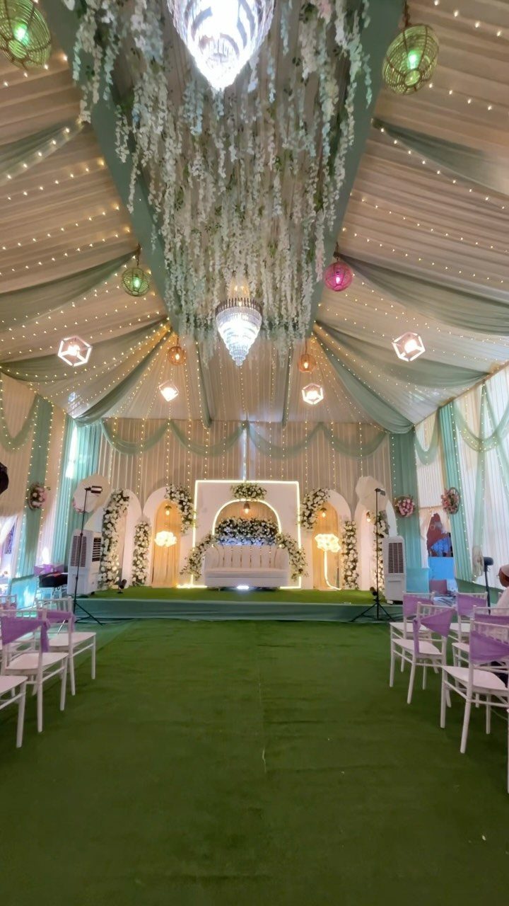 13 Wedding Decor Tent Ideas for 2026 Receptions 13 A decorated wedding tent featuring draped fabric, hanging flowers, chandeliers, and a white stage with green carpeting.