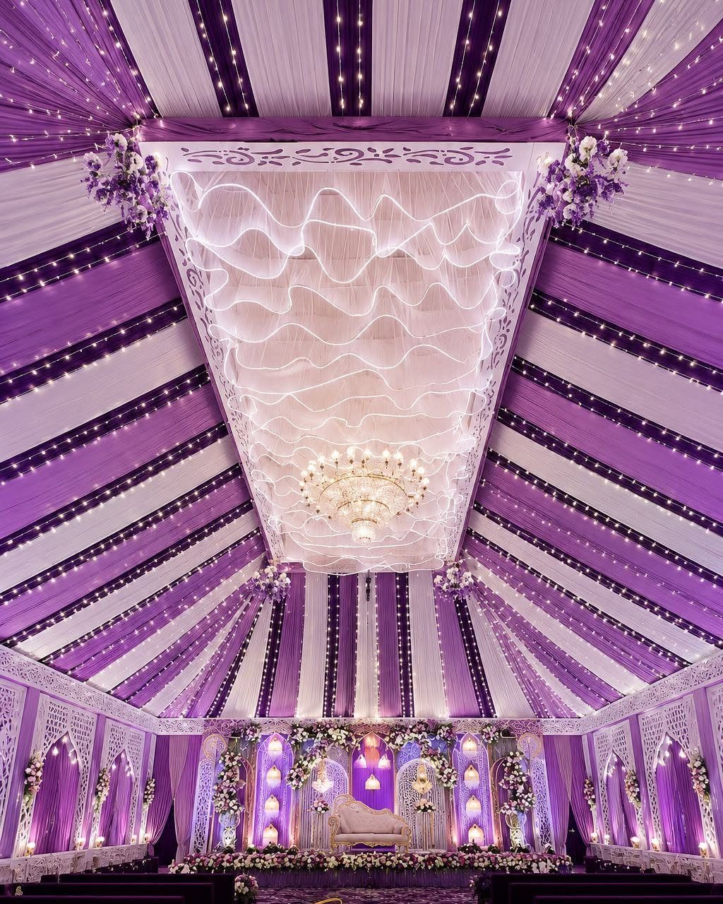 13 Wedding Decor Tent Ideas for 2026 Receptions 1 Purple and white striped tent ceiling with a large white fabric installation and a crystal chandelier.