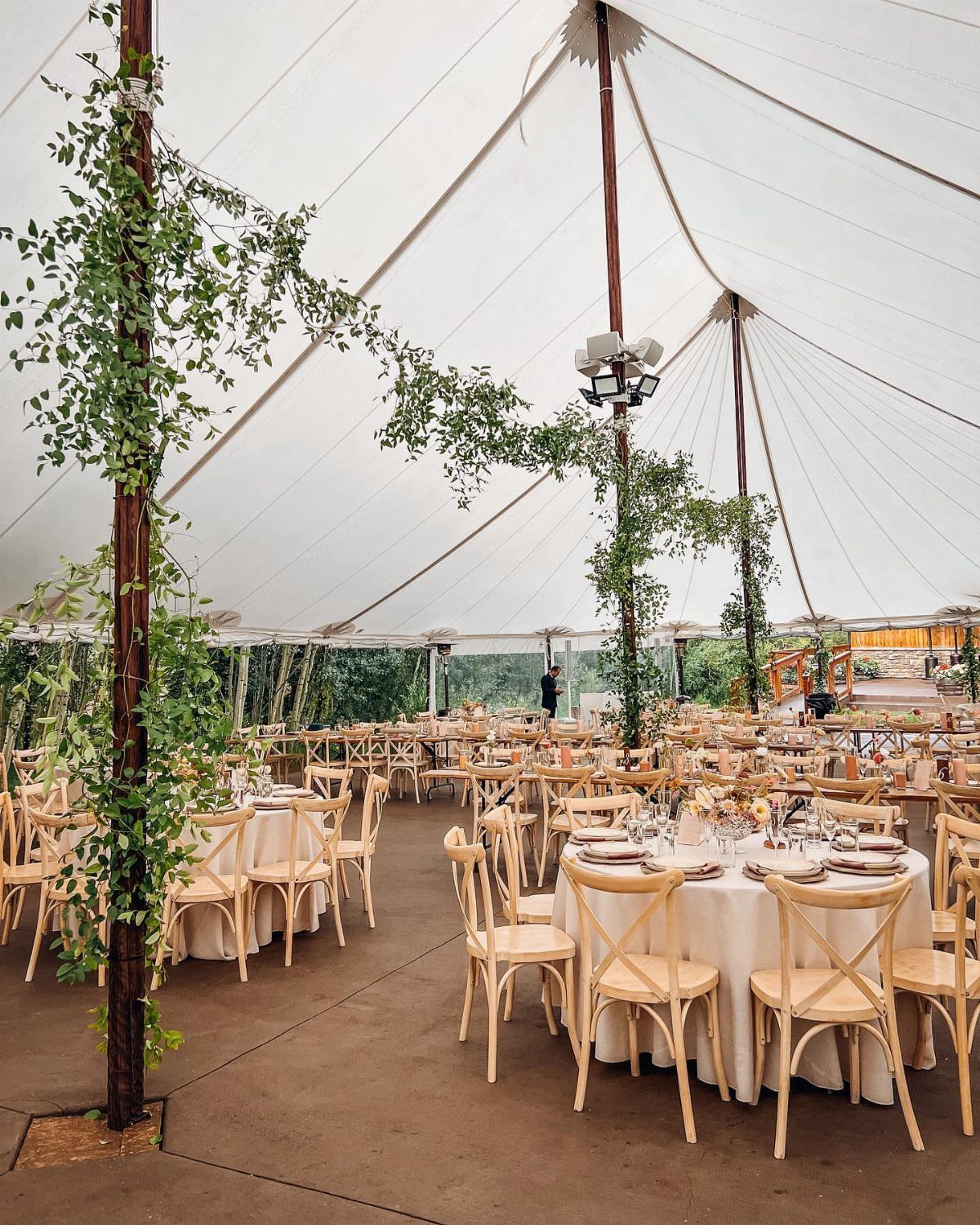 13 Wedding Decor Tent Ideas for 2026 Receptions 3 Wooden tent poles wrapped in green vines inside a decorated wedding reception tent.